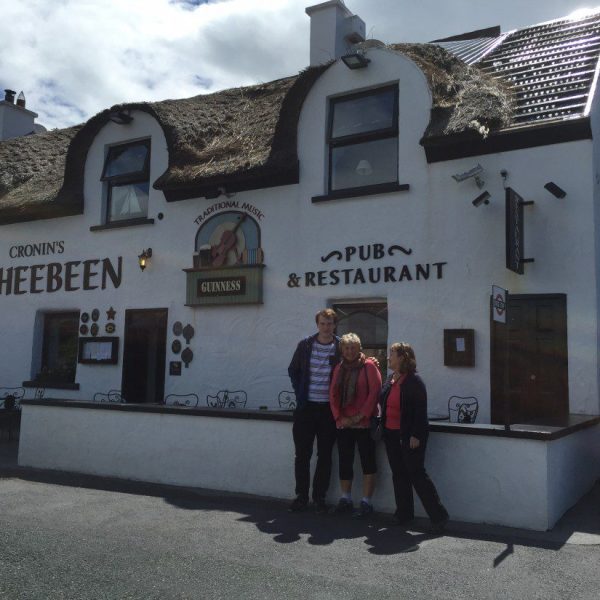 Three people standing in front of cronin's sheebeen pub & restaurant with a thatched roof.