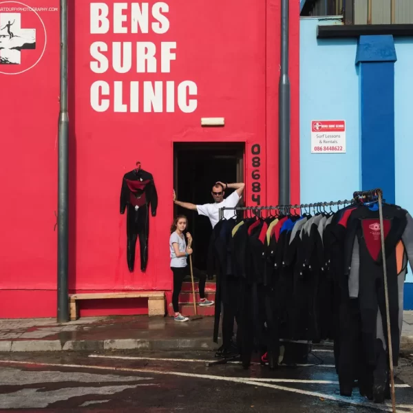 A surf clinic storefront with wetsuits on display and a person with a child at the entrance.