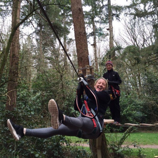 Woman ziplining with an attendant watching from the platform amidst a forest setting.