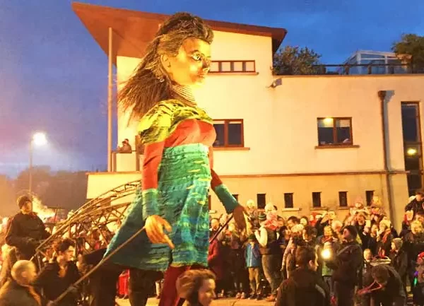 A large puppet character parading through a street event with onlookers gathered around at dusk.