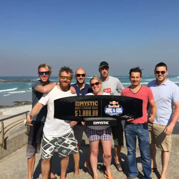 A group of seven people smiling and standing on a sunny beachfront, holding a large novelty check.