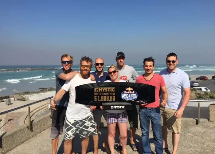 A group of seven people smiling and standing on a sunny beachfront, holding a large novelty check.