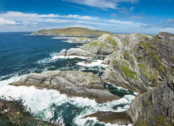 Rugged coastal cliffs against a backdrop of the ocean with an island in the distance under a partly cloudy sky.