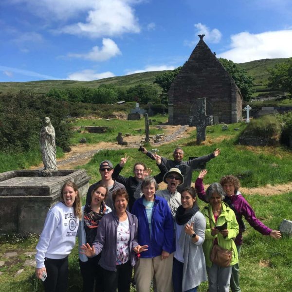 Group of tourists posing for a photo in front of an old stone chapel and graveyard on a sunny day.