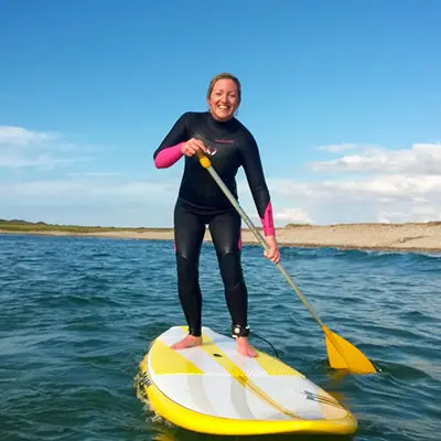 Woman in a wetsuit smiling while stand-up paddleboarding on calm water.