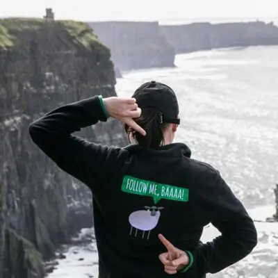 Person wearing a hoodie with a sheep graphic and text "follow me, baaaa" gesturing towards cliffs by the sea.