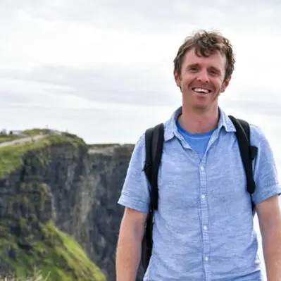 A smiling man with a backpack standing in front of a cliff with a cloudy sky in the background.