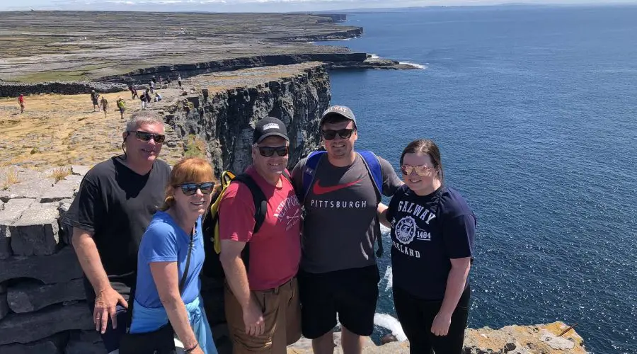 Group of five tourists smiling for a photo with the scenic backdrop of coastal cliffs under a clear blue sky.