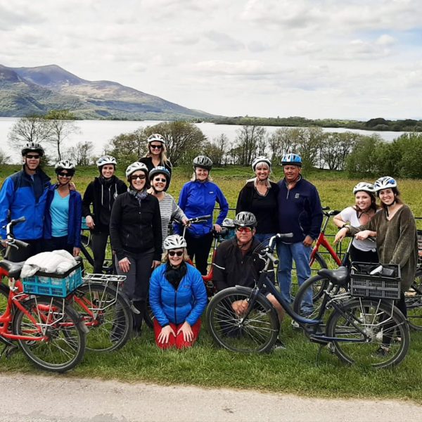 A group of cyclists posing with their bicycles in front of a scenic lake and mountain backdrop.