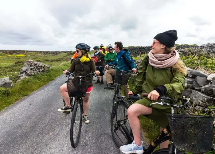 A group of cyclists on one of our small-group Ireland tours makes a stop on a scenic road on the Aran Islands. The sky is overcast, and the landscape looks rugged, with an old drystone wall to one side and long, wild grass growing beside the narrow road.