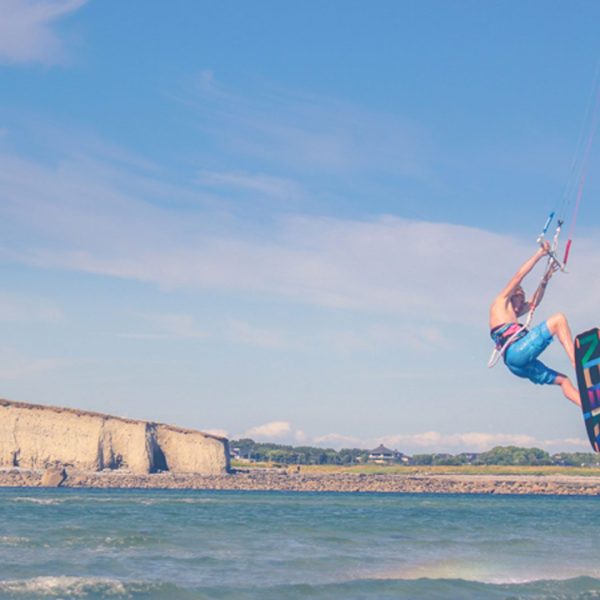 A kite surfer performing a jump over water near a rocky coastline under a clear blue sky.