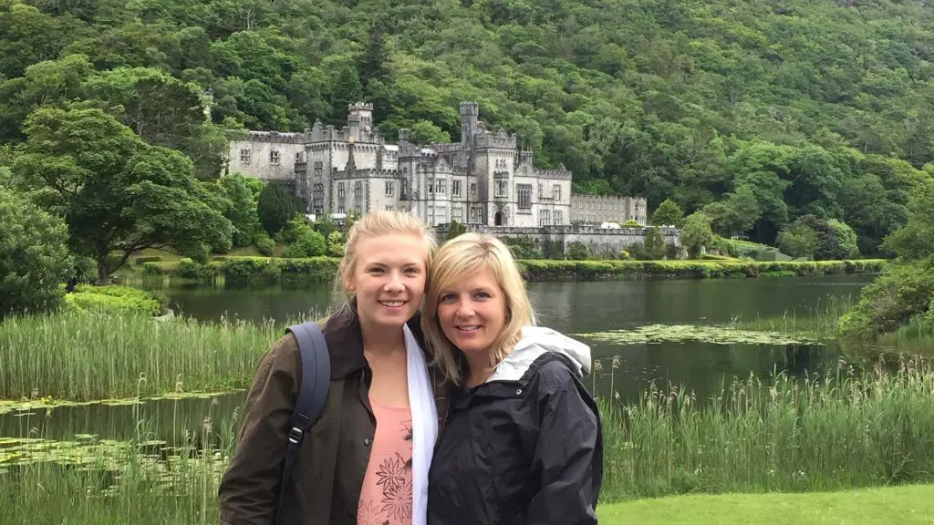 Two women smiling in front of the historic and beautiful Kylemore Abbey in Connemara, County Galway. The abbey overlooks a lake and sits nestled in lush green surroundings, including a forested hillside, the grass banks of the lake, and reeds growing out of the water.