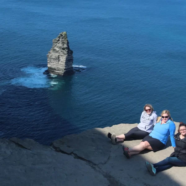 Three people sitting at the edge of a cliff overlooking the sea with a rock formation visible in the water.