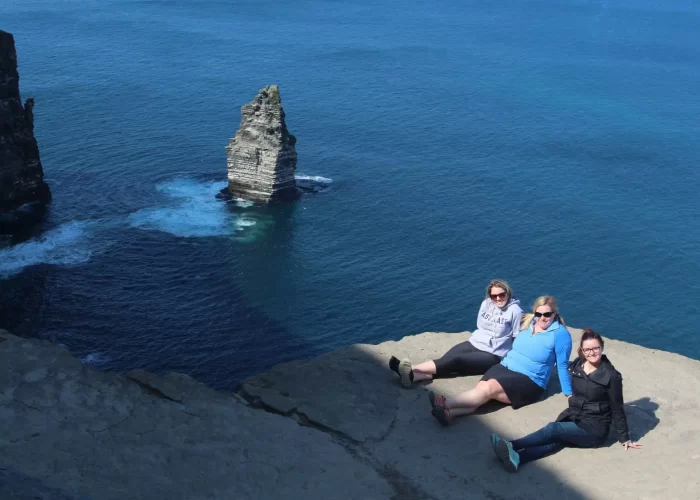 Three people sitting at the edge of a cliff overlooking the sea with a rock formation visible in the water.