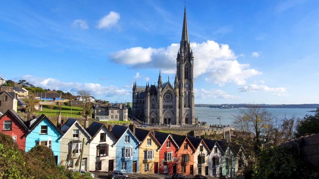The grand St Colmans Cathedral in Cobh, County Cork, rises majestically into a blue, partly cloudy sky. The water of Cork Harbour is visible behind the cathedral, while a row of colorful terraced houses with pitched roofs is in the foreground in front of the cathedral. The houses are on a hill as they stand on a steep gradient.