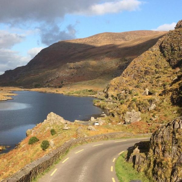 A winding road beside a tranquil lake with rocky terrain and hills under a partly cloudy sky.