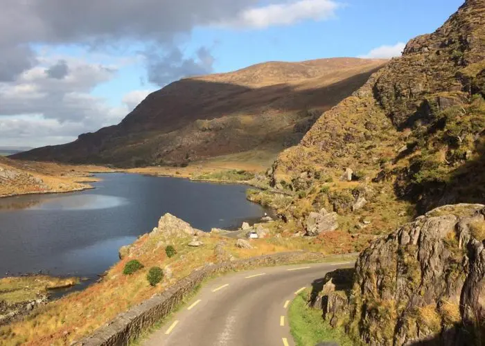 A winding road beside a tranquil lake with rocky terrain and hills under a partly cloudy sky.