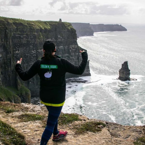 Person standing at the edge of the cliffs of moher, giving a thumbs-up to the scenic view.