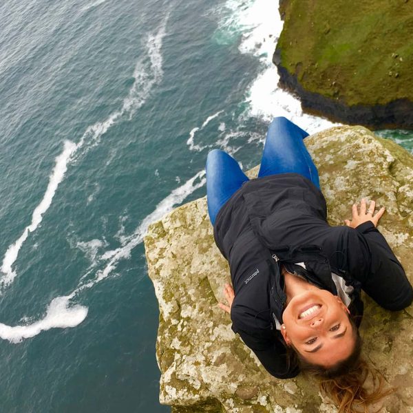 Woman lying on her back on a cliff edge, smiling at the camera with waves breaking against the coastline below.