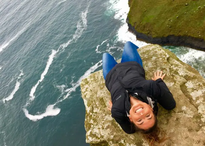 A woman smiling, lying on her back on a cliff edge, looks up and smiles for the camera. The Atlantic Ocean rolls in below her and there is a smaller green clifftop in the background. This sense of adventurous fun on the Wild Atlantic Way is what Overland’s small-group tours are all about.