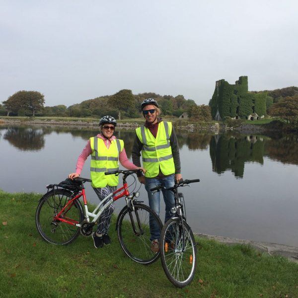 Two cyclists wearing high-visibility vests pose with their bikes by a calm lake with a castle ruin in the background.