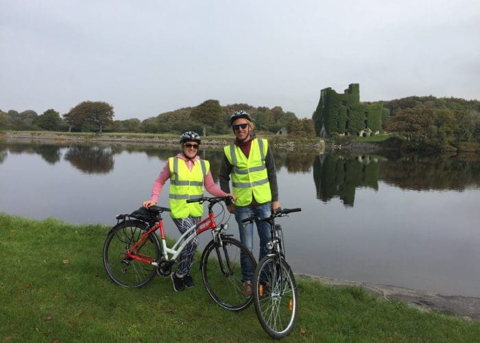 Two cyclists wearing high-visibility vests pose with their bikes by a calm lake with a castle ruin in the background.