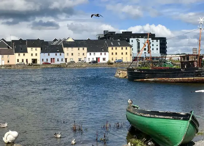 Colorful houses line the waterfront with birds in the foreground and a green boat beached nearby, under a partly cloudy sky.