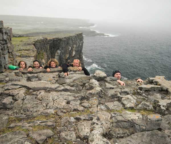 Group of people peeking over a stone wall with a foggy cliffside coastline in the background.