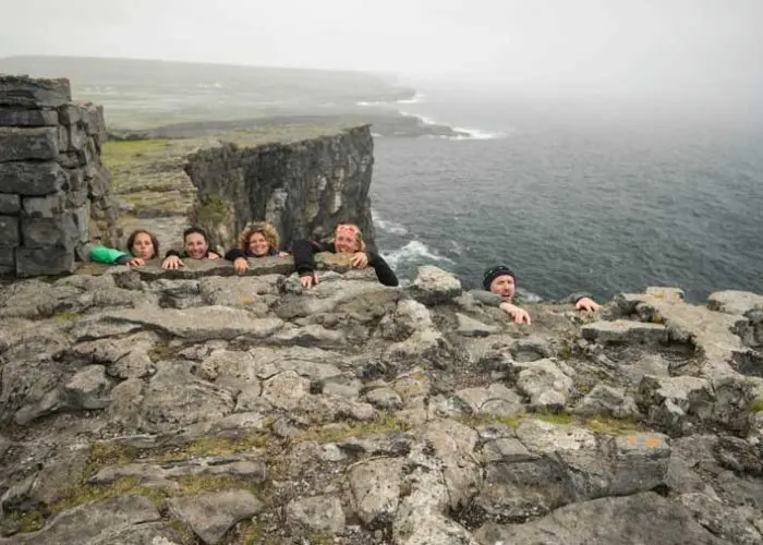 Group of people peeking over a stone wall with a foggy cliffside coastline in the background.