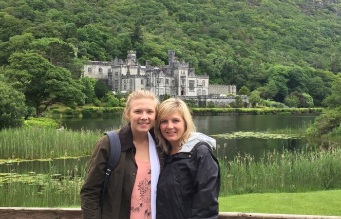 Two people smiling in front of an historic building by a lake with lush greenery in the background in Ireland.