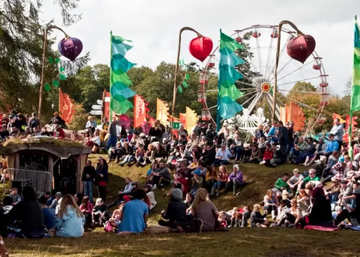 Outdoor festival with attendees sitting on the grass enjoying performances near colorful flags and amusement rides.