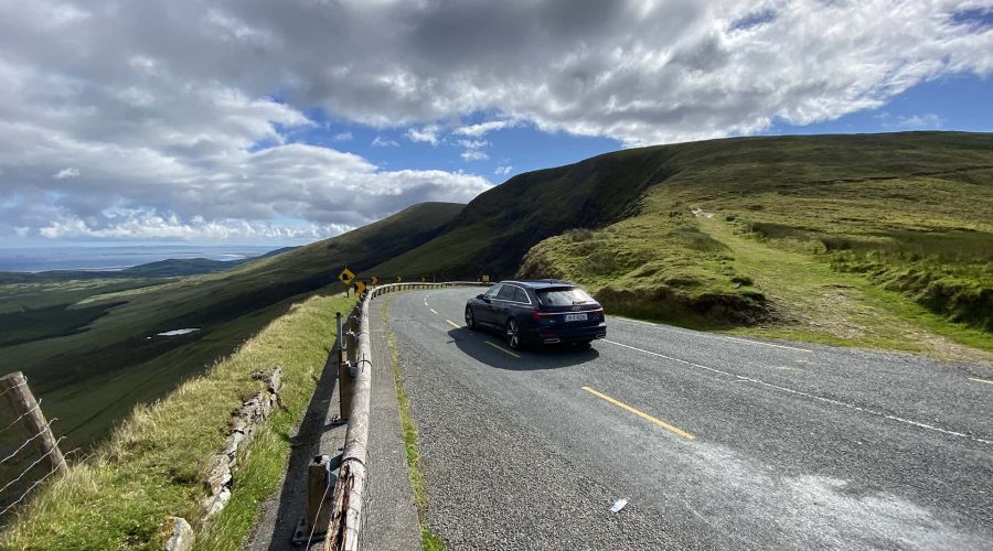 Car driving on a winding mountain road with scenic hills and cloudy sky in the background.