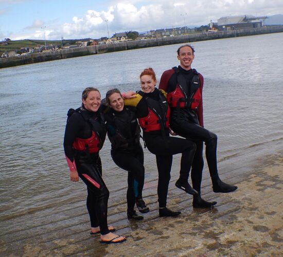 Group of four people in wetsuits and life jackets smiling on a beach in Ireland, presumably after or before watersports.