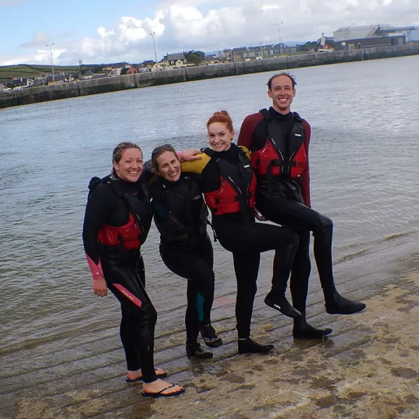 Four people in wetsuits and life jackets posing on a sandy beach with water and buildings in the background.