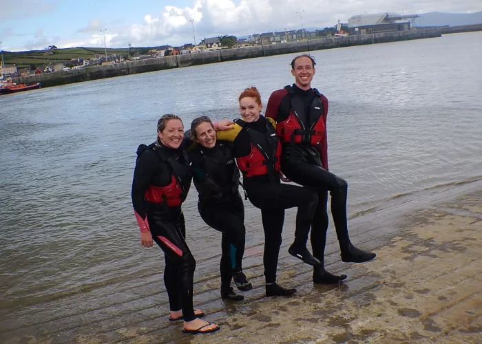 Four people in wetsuits and life jackets posing on a sandy beach with water and buildings in the background.