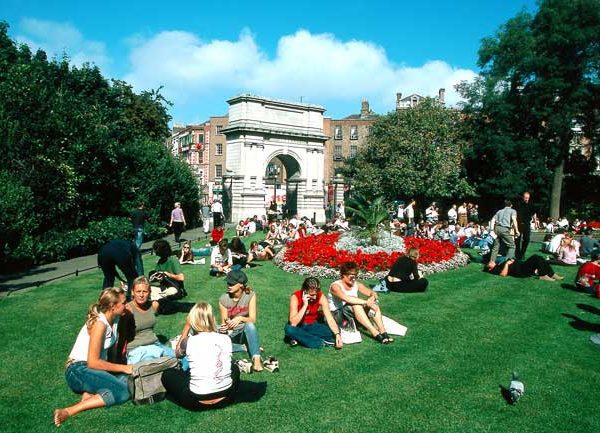 Groups of people enjoying a sunny day on the grass in a city park with a monument in the background.