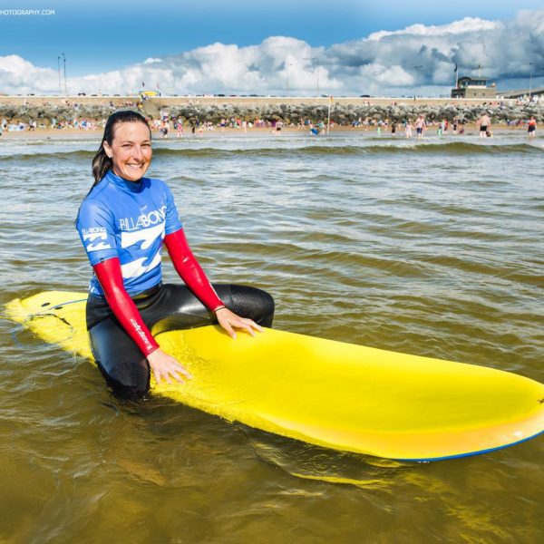 A smiling surfer in a blue wetsuit sitting on a yellow surfboard near the shore, with a crowd and buildings in the background.