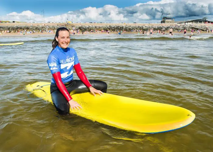 A smiling surfer in a blue wetsuit sitting on a yellow surfboard near the shore, with a crowd and buildings in the background.