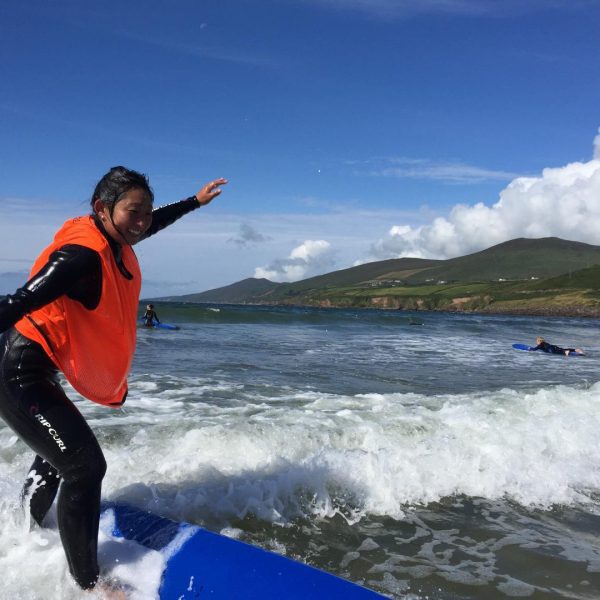 A person in a wetsuit balancing on a surfboard at the beach with hills in the background.