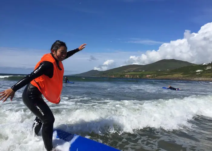 A person in a wetsuit balancing on a surfboard at the beach with hills in the background.