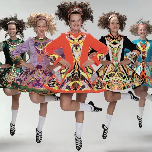 Five irish dancers in colorful dresses performing a jump with their arms held rigidly by their sides.