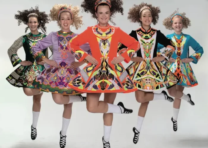 Five irish dancers in colorful dresses performing a jump with their arms held rigidly by their sides.