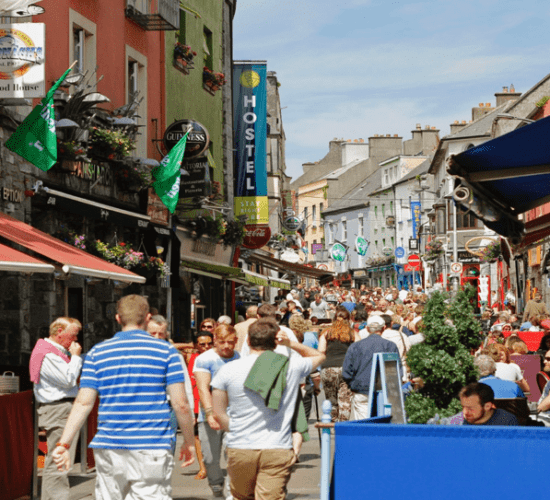 Crowded street in a sunny urban setting with pedestrians, outdoor seating, hanging flower baskets, and small group tours.