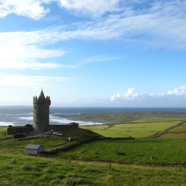 An ancient stone tower overlooking a coastal landscape under a bright blue sky with scattered clouds.