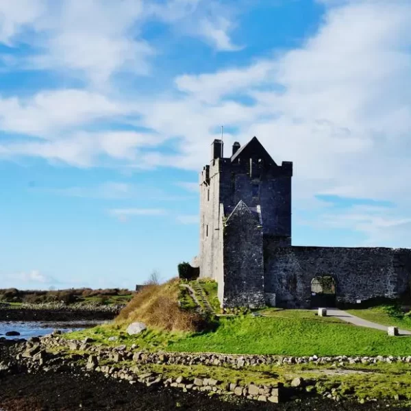 An ancient stone castle stands on a grassy mound under a blue sky with scattered clouds.