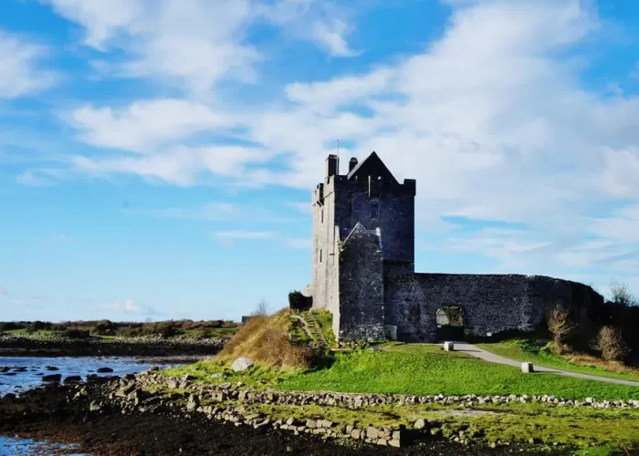 An ancient stone castle stands on a grassy mound under a blue sky with scattered clouds.