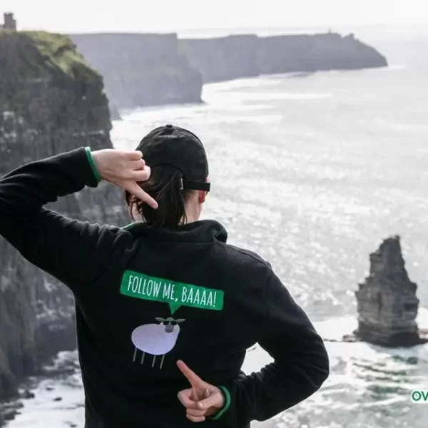 A person wearing a hoodie with a sheep design and the text "follow me, baaa!" is pointing towards the ocean while standing at the edge of a cliff.