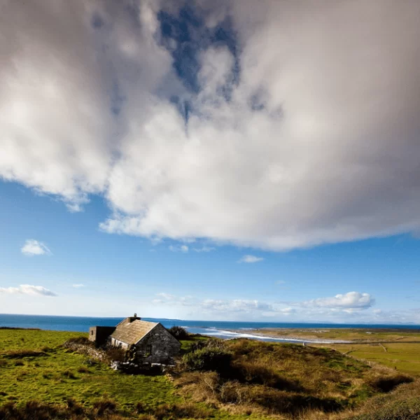 A solitary stone house stands on a grassy field under a vast sky with scattered clouds.