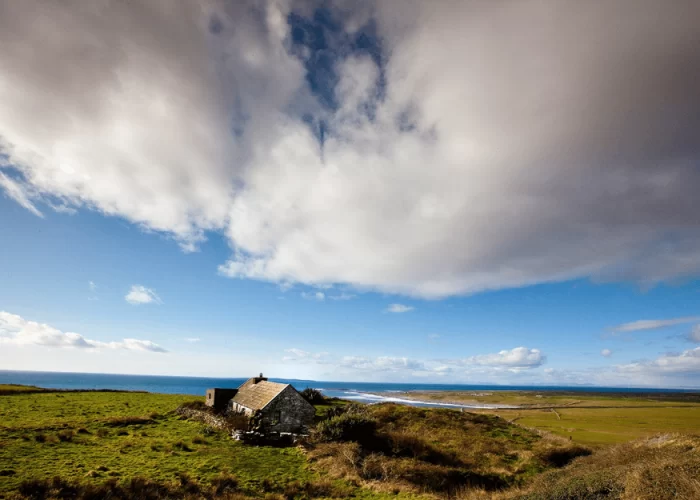 A solitary stone house stands on a grassy field under a vast sky with scattered clouds.