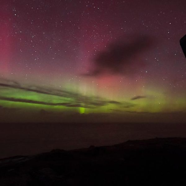 Aurora borealis illuminates the night sky with green and pink hues over a coastal landscape with a silhouette of a structure on the right.